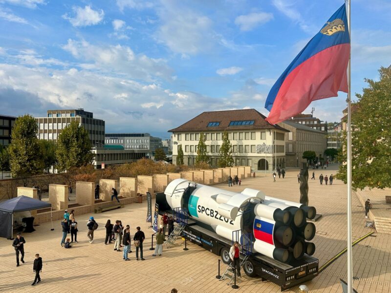 Raketenförmige VR-Anlage SpaceBuzz auf dem Peter-Kaiser-Platz in Vaduz, daneben Besucherinnen und Besucher und die Fahne Liechtensteins